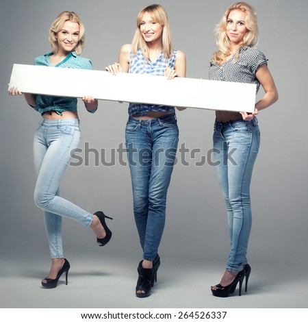 Three beautiful young woman posing in fashionable jeans, holding empty board. Blonde girls. Studio photo.