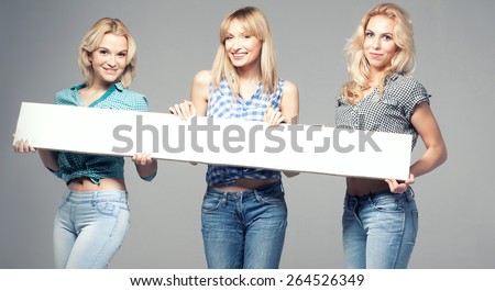 Three beautiful young woman posing in fashionable jeans, holding empty board. Blonde girls. Studio photo.