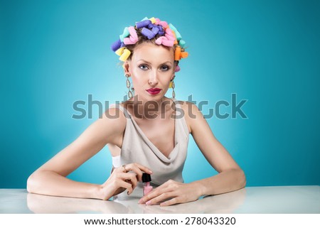 Beauty portrait of attractive woman posing with nail polish and with curlers on her head. Blue background.