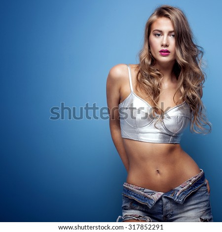 Sexy beautiful young woman posing on blue background, looking at camera. Girl in short jeans and silver fashionable top. Studio shot.