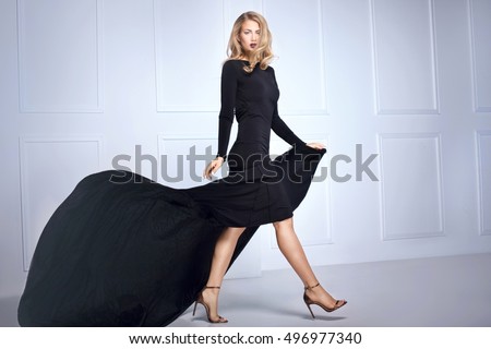 Beautiful elegant blonde woman posing in black maxi dress, looking at camera. Indoor shot.
