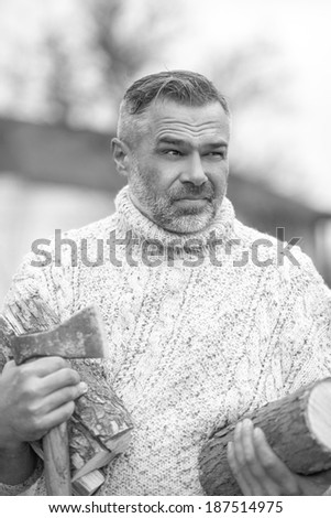 Black and white portrait od handsome man wearing sweater holding axe and wooden logs for firewood