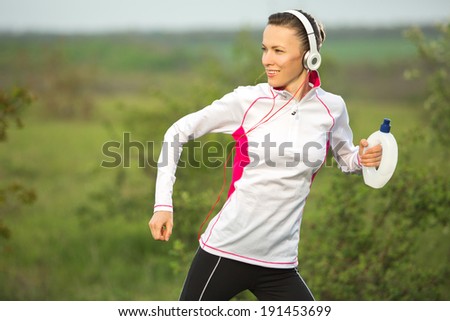 Running woman - female runner listening to music on smart phone hold water bottle outdoor in motion. Sporty beautiful woman jogging and listening to training music on smartphone
