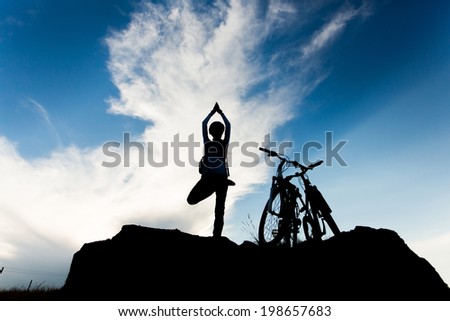 Silhouette young woman practicing yoga, relaxing after riding bikes high in mountain over cloudy sky background