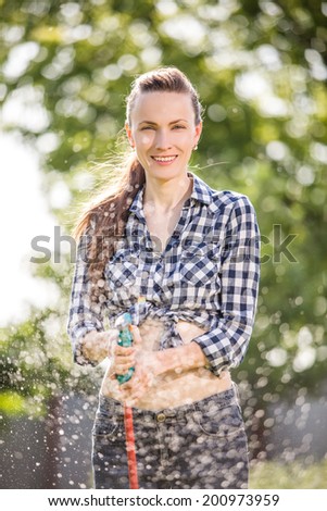 Beautiful young woman having fun in summer garden with garden hose splashing summer rain. soft backlight