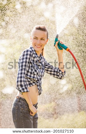 Beautiful young woman having fun in summer garden with garden hose splashing summer rain. soft backlight