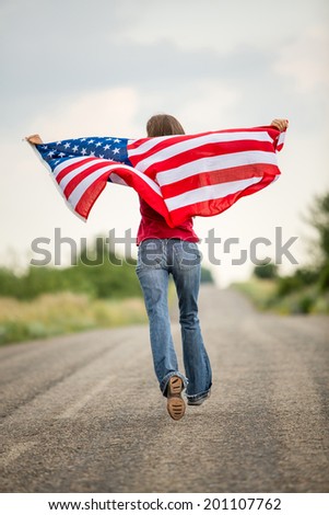 Patriotic young woman with the American flag held in her outstretched hands running along the road in motion. focus on flag
