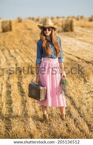 Pretty young woman hitchhiking along a road and relaxing on beautiful field with hay stacks.