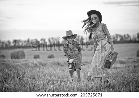 Black and white image of funny mother and a son in cowboy hats playing in travelers with a toy horse outdoors. in motion, focus on boy