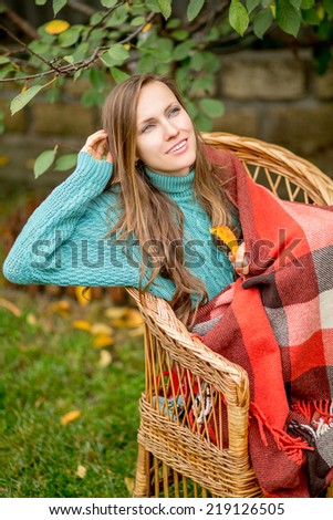 Beautiful woman in the garden wrapped in a plaid blanket sitting in armchair. Sweater autumn woman