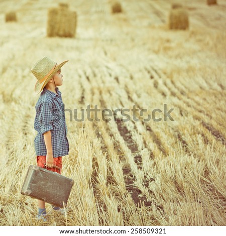 Small traveler boy standing dreaming on summer field wearing cowboy hat and holding old  suitcase. toned old vintage image