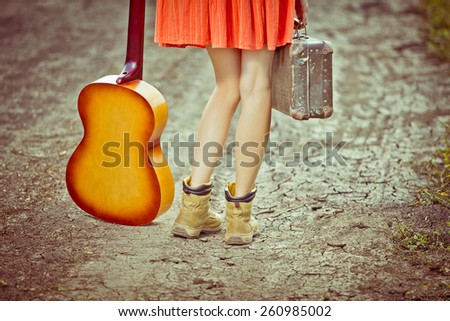 Legs of a woman tourist with suitcase and guitar standing on rural road. toned vintage image, vignette,  copy space