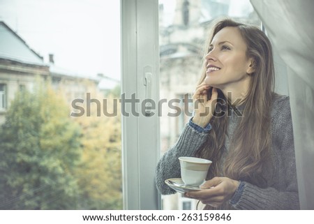 beautiful woman drinking coffee by the window on a rainy cloudy day, focus on face, toned filter image