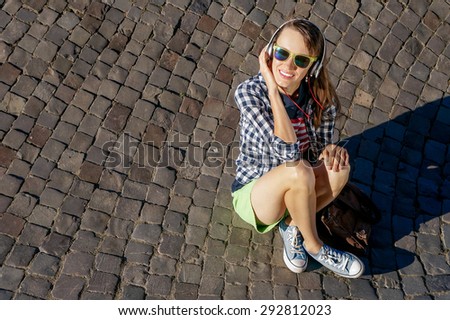 Top view young woman sitting on paving stones enjoying the sunshine and listening music, tourist girl in bright glasses relaxing outdoors