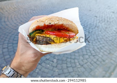 Cheeseburger with tomato and pickles on a sesame seed bun in man hand over grey pavement. view from above