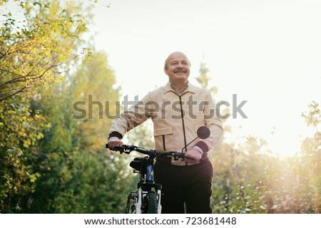 Senior caucasian man go for a walk with bike in countryside. Green trees on background. He is happy and active.