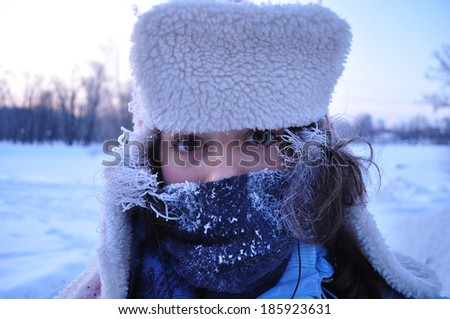 A face of a young girl with frozen hair and scarf with snowy forest at the background