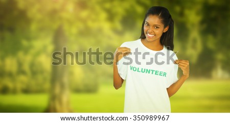Young woman wearing volunteer tshirt and pointing to it against trees and meadow