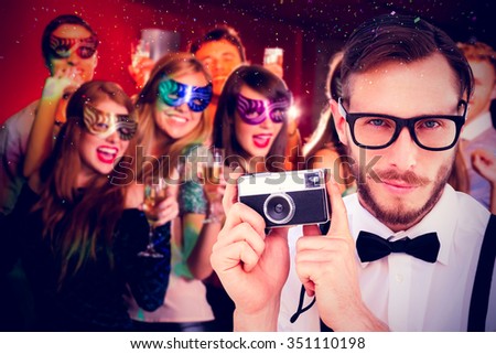 Geeky hipster holding a retro camera against friends in masquerade masks drinking champagne