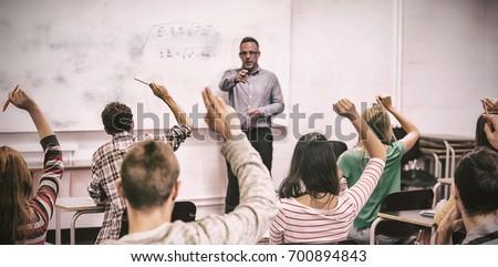 Rear view of students with hands raised with teacher in classroom