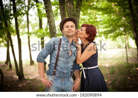 A beautiful young woman whispers to an handsome young man's ear and he smiles, in the wood, with a beautiful sun light, in a stylish and vintage-like mood.
