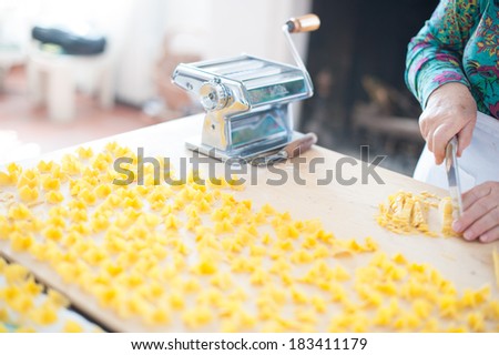 A traditional pasta machine on a wooden cutting board close to drying home made pasta and woman\'s hands cutting a fresh pasta sheet.