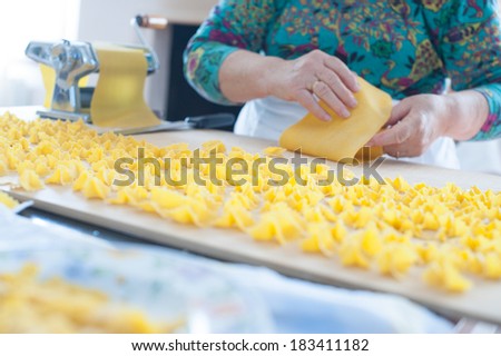 Woman\'s hands roll up a fresh pasta sheet on a wooden cutting board full of drying home-made pasta pieces and close to a traditional metal pasta machine.
