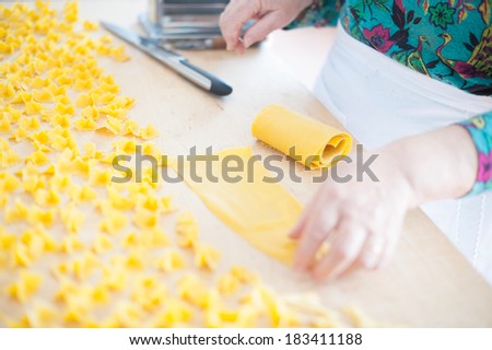 Woman\'s hands hold a fresh pasta sheet on a wooden cutting board close to home made drying pasta pieces.