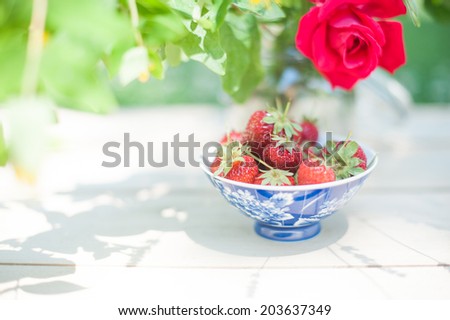 A pot full of strawberries under the shadow of a vase of flowers, in a shabby chic mood.
