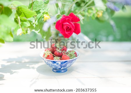 A colorful pot of strawberries under the shadow of a vase full of flowers, in a shabby chic mood.