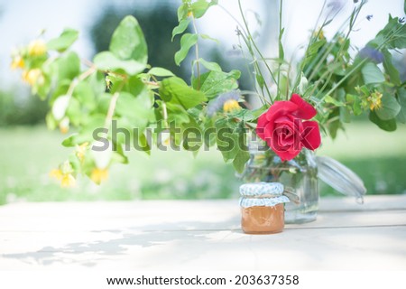 A tiny pot of marmalade under the shadow of a vase full of flowers, in a shabby chic mood.