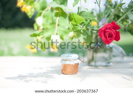 A tiny pot of home made marmalade under the shadow of a vase full of flowers, in a shabby chic mood.