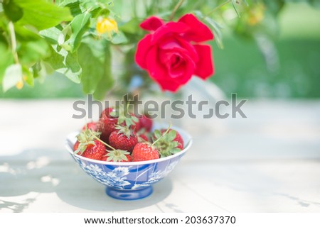 A colorful pot with strawberries under the shadow of a vase full of flowers, in a shabby chic mood.