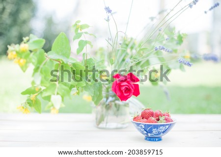 Front view of a cup full of strawberries and a vase of flowers, in a shabby chic mood.