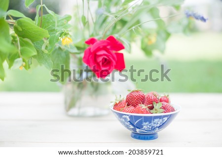 Close up of a cup full of strawberries and a vase of flowers, in a shabby chic vintage mood.