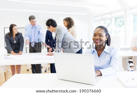 African ethnic woman smiling at the camera while working on her laptop with business meeting as the background.