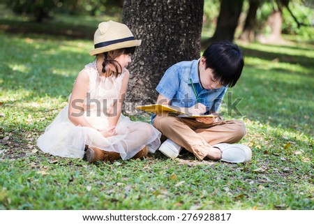 Little asian boy use pencil writing on notebook for writing book with smiling face in the park and younger sister looking to her brother