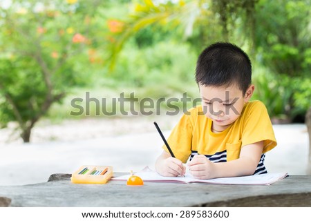 Little asian boy use pencil writing on notebook for writing book with smiling face on wooden table in the park