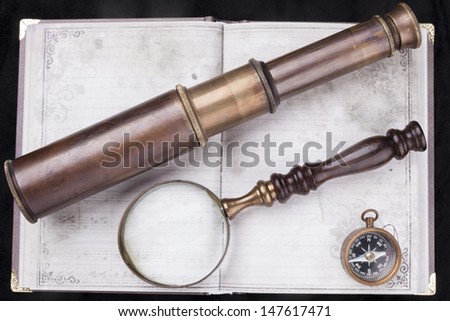 Old telescope with magnifying glass and a compass on an old notebook with black background