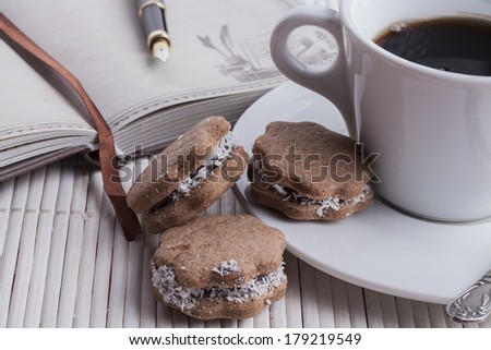 brown cookies with coffee in a white mug and silver spoon with fountain pen and old notebook on white wood background