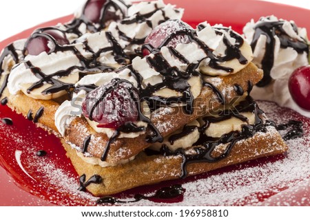 Belgian waffle with chocolate syrup whipped cream and banana slice with red cherry on red plate and sugar powder snow