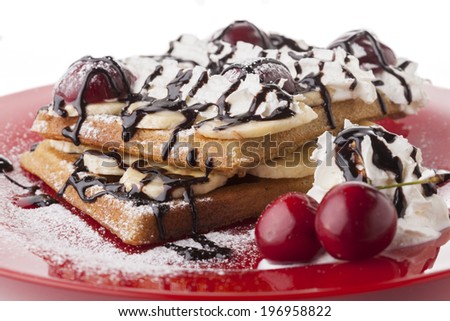 Belgian waffle with chocolate syrup whipped cream and banana slice with red cherry on red plate and sugar powder snow