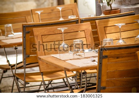 Photo presents old town outdoor restaurant, visible wooden table and chairs, glasses and wine glasses put upside-down on the table mats.
