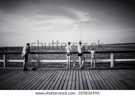 Photo presenting 4 people - 3 men and child, All standing backwards, fishing with fishing rods, on pier. People are unrecognizable, model release not necessary. Black and white photo.