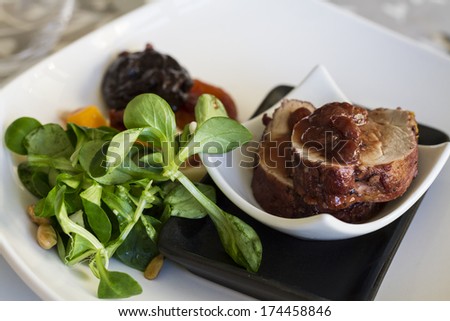 Small portions of pork tenderloin steaks with lamb\'s lettuce salad, garnish accompanied by dried apricot and raisins on small black and white plates over wooden table