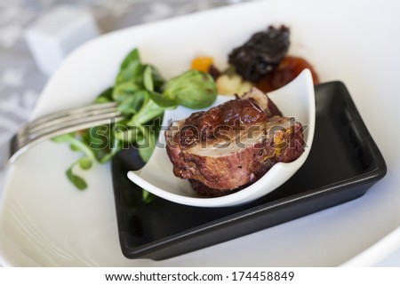 Small portions of pork tenderloin steaks with lamb\'s lettuce salad, garnish accompanied by dried apricot and raisins on small black and white plates over wooden table