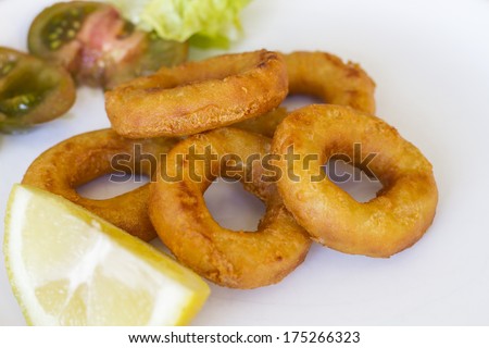 Five breaded fried calamari rings with lemon slice, accompanied by tomato and lettuce on white plate over wooden table