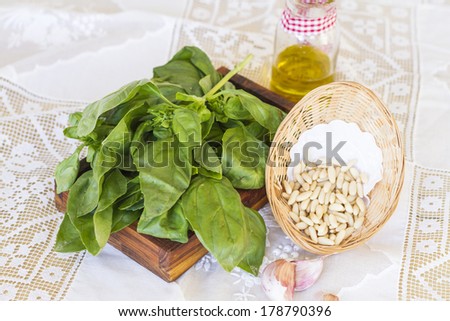 Bunch of basil on wooden stand and basket with pine nuts on white tablecloth, decorated olive oil jar and some garlics.