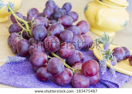 Still life composed of bunch of grapes on purple napkin and wooden table, with different yelow elements and decorated with violet flowers.
