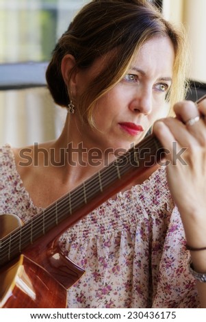 Attractive white woman in her forties playing guitar and looking pensively out the window and wearing floral shirt and vintage romantic style.